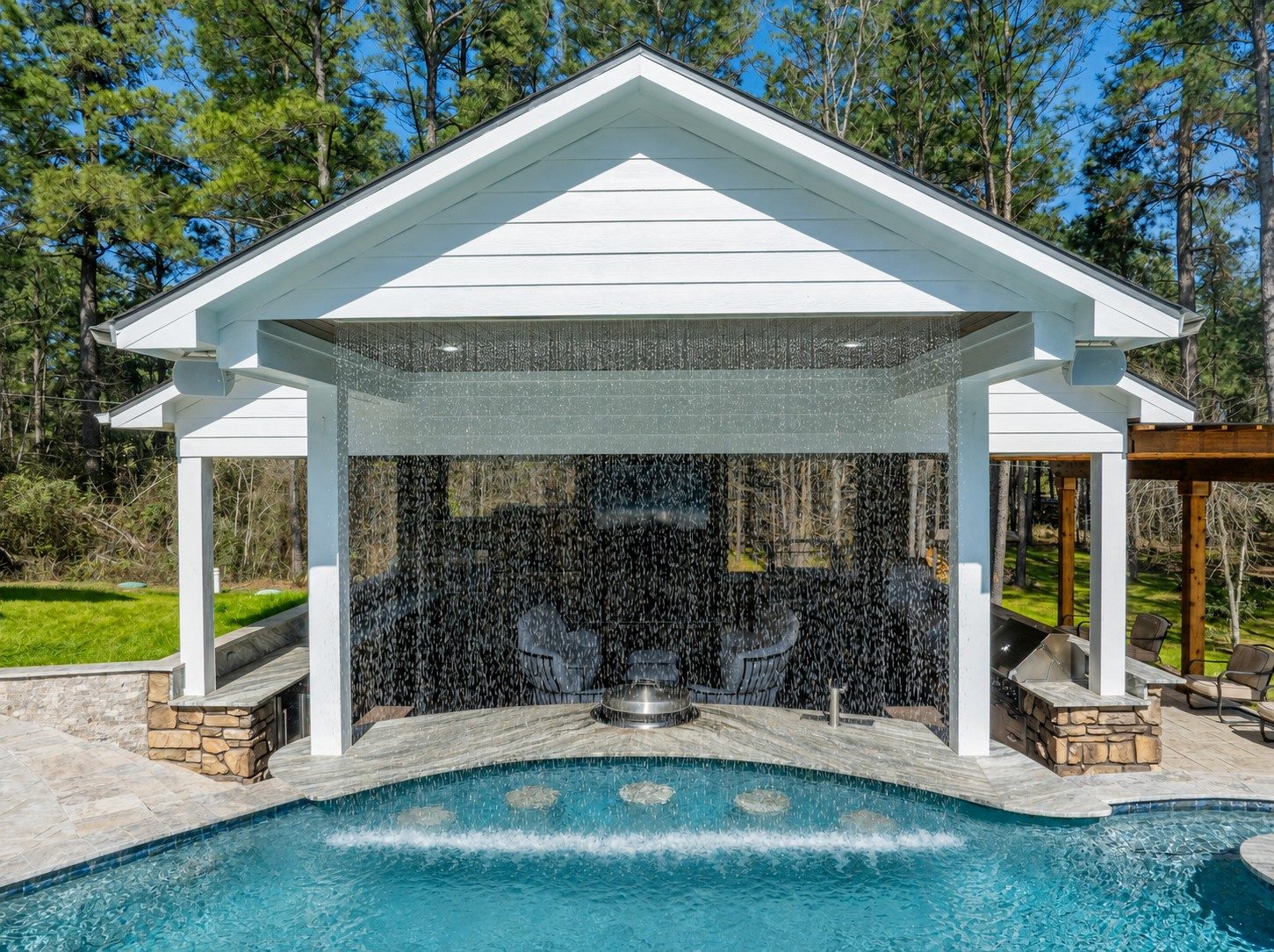 A curtain of water cascades from a white pavilion roof into a freeform pool screening a lounge seating area beside an outdoor kitchen