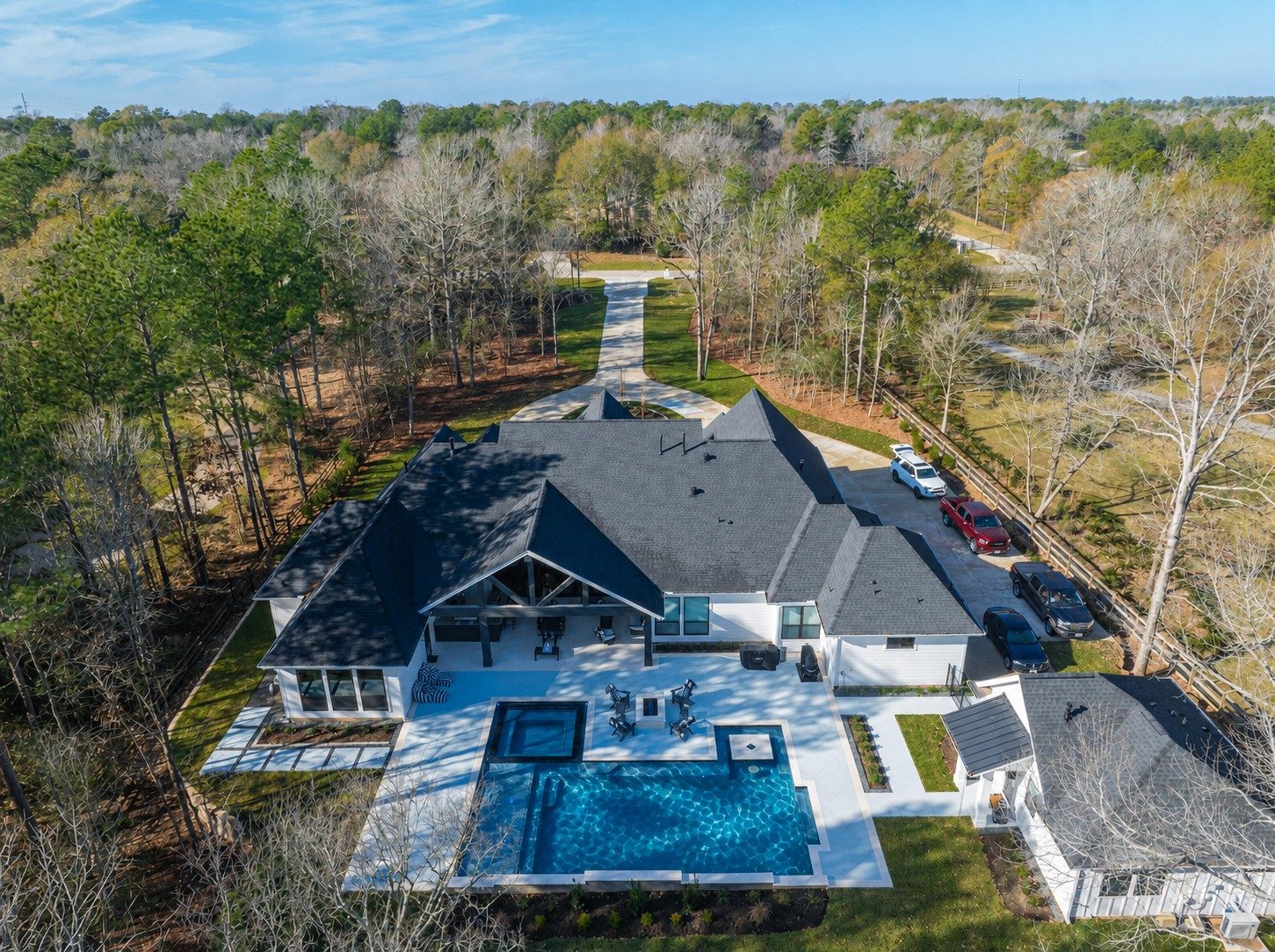 High aerial of the modern farmhouse on a wooded lot with the geometric pool centered in a white deck